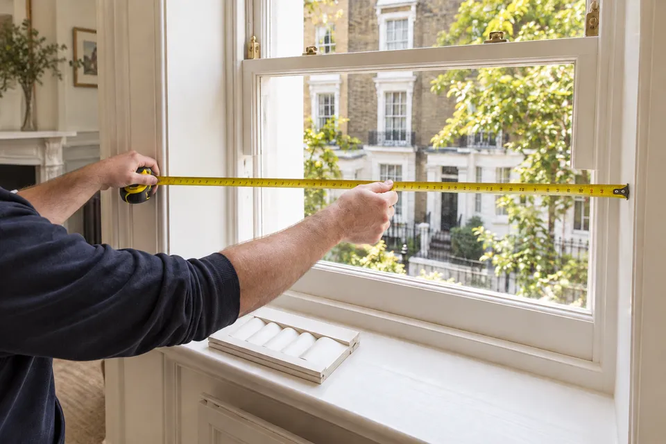 Hands measuring the width of a white sash window recess with a tape measure